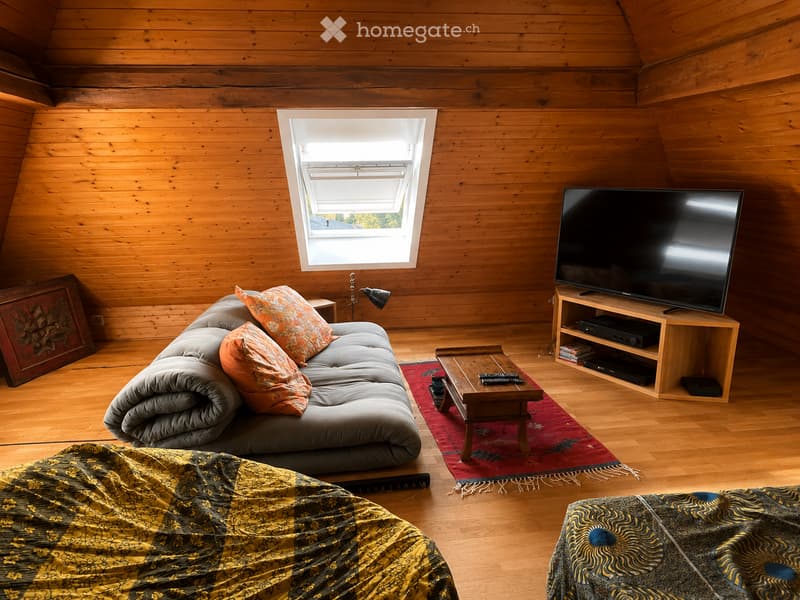 Cozy living room with wooden walls and floor, featuring a grey sofa with orange cushions, a wooden coffee table, a flat-screen TV on a wooden stand, and a floor lamp near the window.
