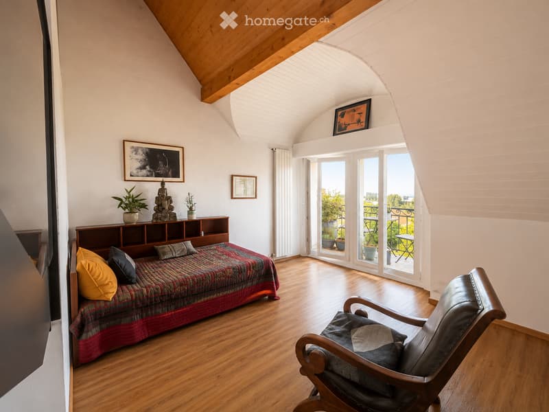 Bright bedroom with wooden floors, slanted wooden ceiling, large windows opening to a balcony, bed with colorful quilt, leather armchair, small table, shelves with decorative items, and framed artwork on walls.