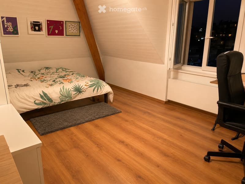 Cozy bedroom with wooden flooring, a bed with a tropical-patterned duvet, a black office chair, and a small rug near the bed.
