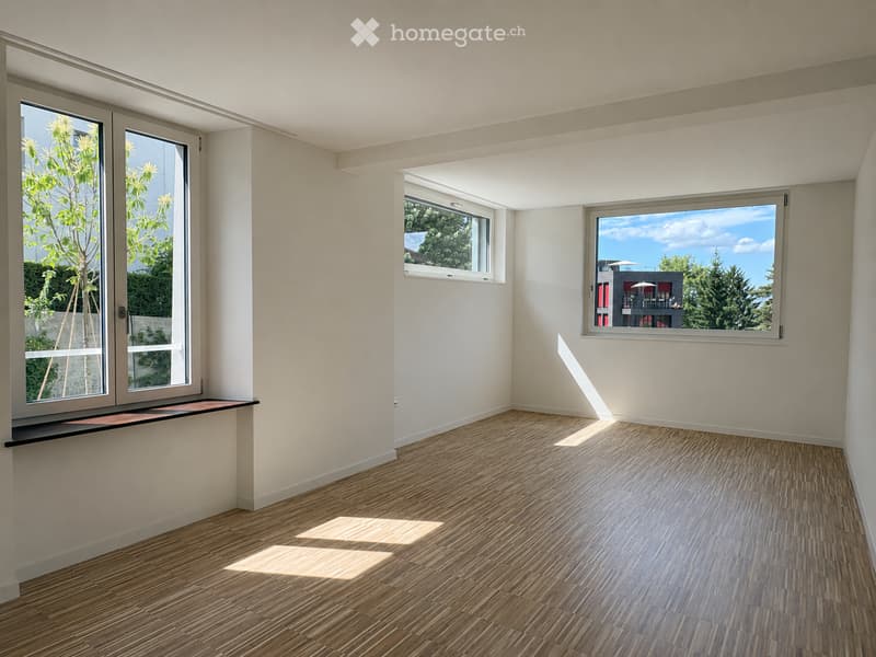 Empty room with light-colored walls, wooden flooring, and multiple windows offering views of greenery and a neighboring building.