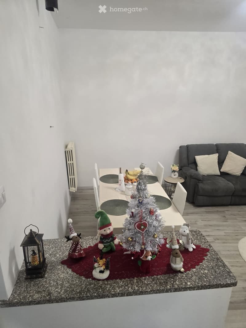 A cozy living and dining area with a dark grey sofa, a dining table with chairs, and a small Christmas tree on the dining table surrounded by festive decorations.