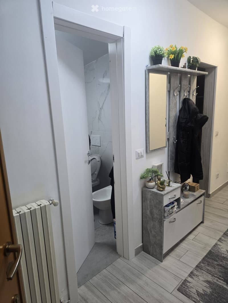 Hallway with wooden flooring, a radiator, a small vanity with a mirror, potted plants, and an open door leading to a bathroom with a toilet and bathtub.