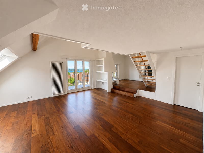 Bright, empty attic room with wooden flooring, white walls, and large windows offering a scenic view. Features a staircase leading to another level and built-in shelving units.