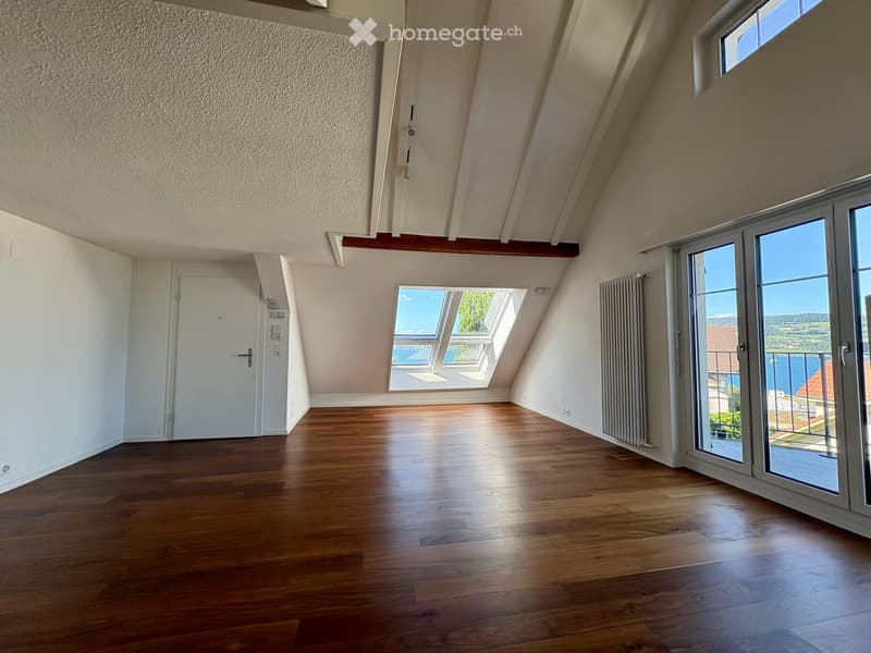Bright, empty attic room with wooden flooring, white walls, and large windows offering a scenic view. The room features a sliding glass door leading to a balcony.