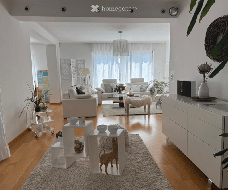 Modern living room with light-colored furniture, white walls, wooden flooring, and large windows with sheer curtains. Features include a white sofa, coffee table, side tables, floor lamp, decorative vases, and plants.
