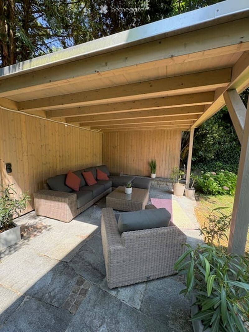 A covered outdoor patio with a grey sofa, coffee table, wicker chair, and several potted plants arranged on a stone floor.
