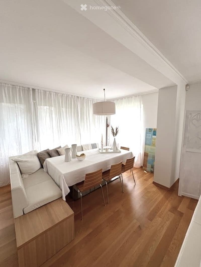 Bright dining area with a white tablecloth-covered dining table, four wooden chairs, a built-in bench seating, and a floor lamp. The room features wooden flooring and large windows with white curtains.