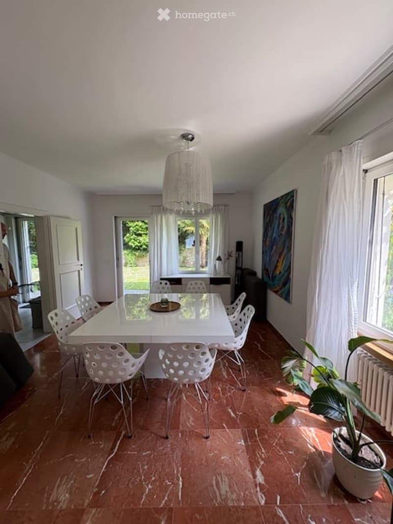 Bright dining room with a white dining table and six white chairs, a large potted plant, a painting on the wall, and a chandelier hanging from the ceiling. The room has large windows with white curtains and a wooden floor.