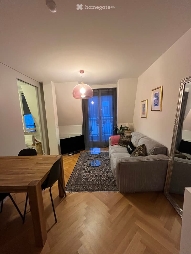 A modern living room with wooden flooring, a grey sofa with patterned cushions, a glass coffee table, a wooden dining table with chairs, a large mirror, and a hanging lamp.