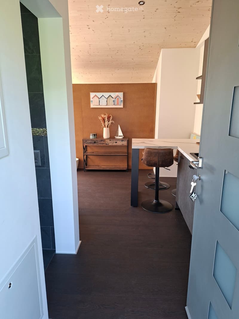 A modern, open-concept kitchen and dining area with dark wood flooring, white cabinets, and a central island with a bar stool. A small wooden table with a vase, sailboat model, and picture frame is visible.