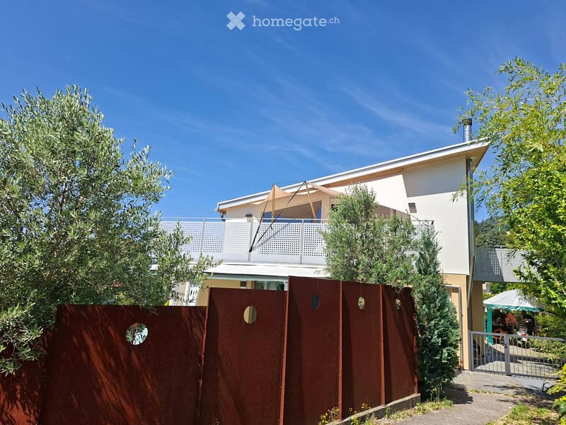 Two-story modern house with white walls, a balcony, and surrounding greenery including olive trees and shrubs.