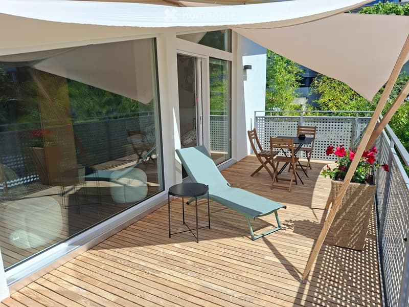 Wooden deck with sun lounger, small table, chairs, and flower pot, with a large umbrella providing shade.