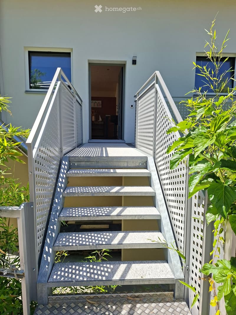 Exterior staircase leading to a raised ground floor entrance, with metal railings and steps, surrounded by greenery.