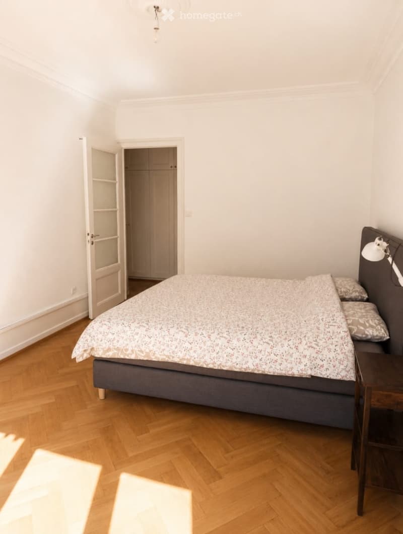 A modern bedroom with a wooden floor, a bed with a patterned duvet, a nightstand with a lamp, and a closet in the background.