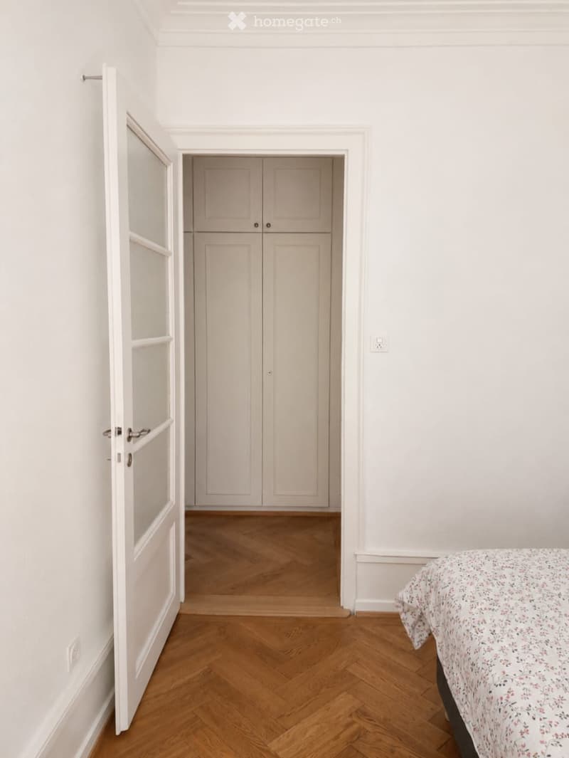 A bedroom with herringbone wooden flooring, white walls, and a bed with a floral-patterned duvet visible in the foreground. A closet with double doors is visible in the background.