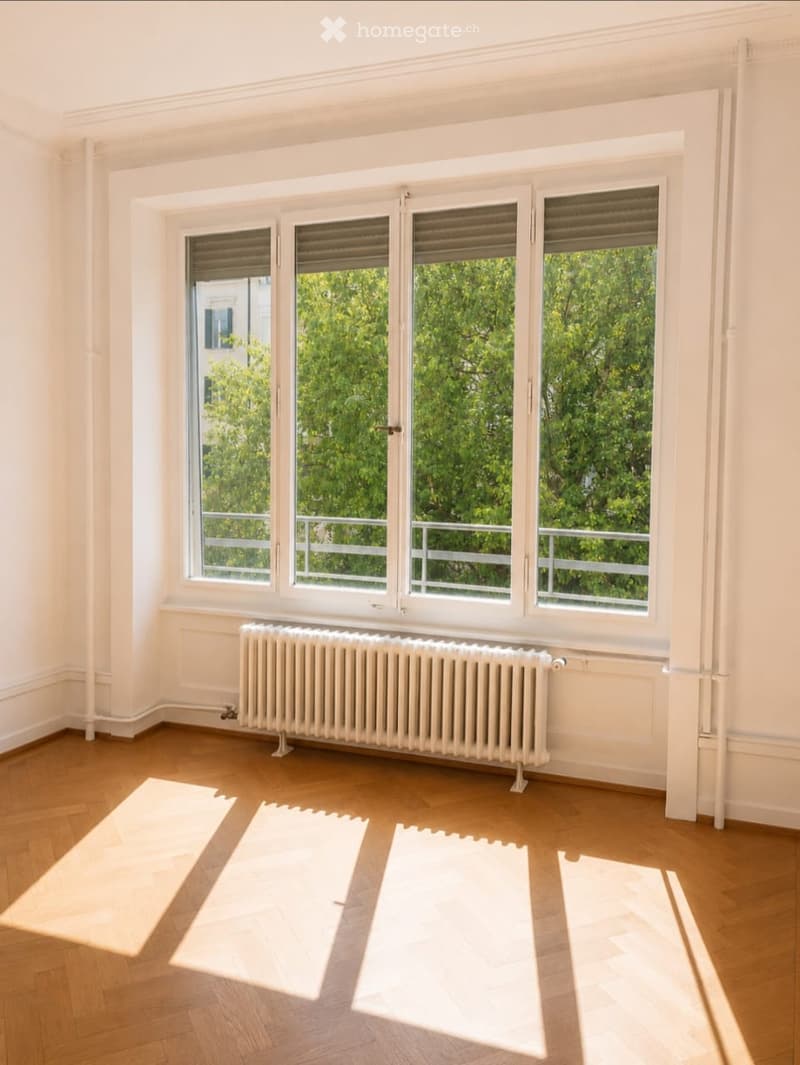 Empty room with wooden flooring, white walls, large windows with blinds, radiator, and natural light from the windows.