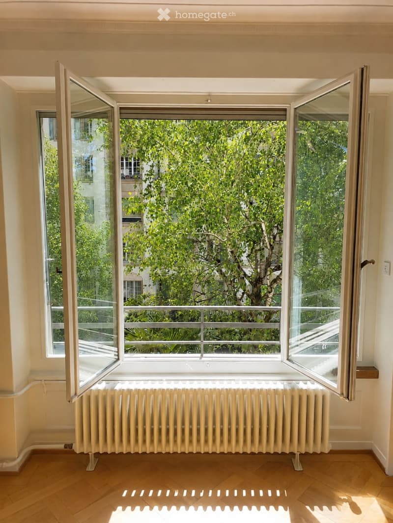 Empty room with wooden flooring, large open windows, radiator, and a view of greenery outside.