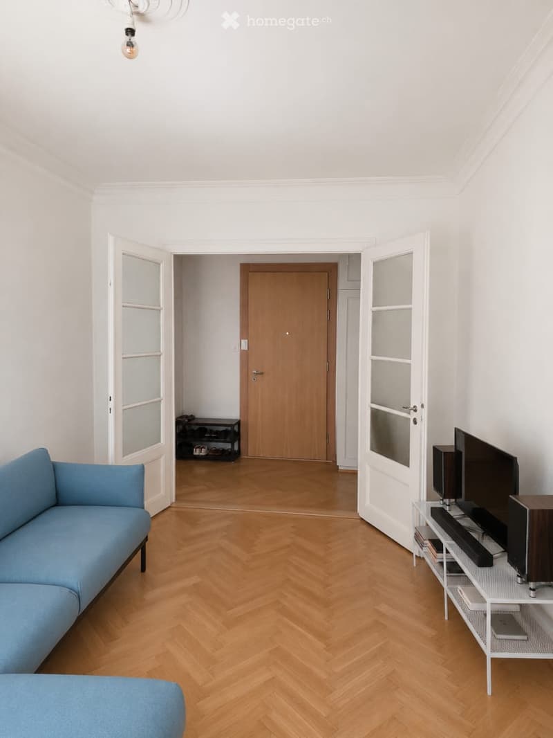 Bright living room with herringbone wooden flooring, blue sofa, TV stand, and a shelf with books and electronic devices.