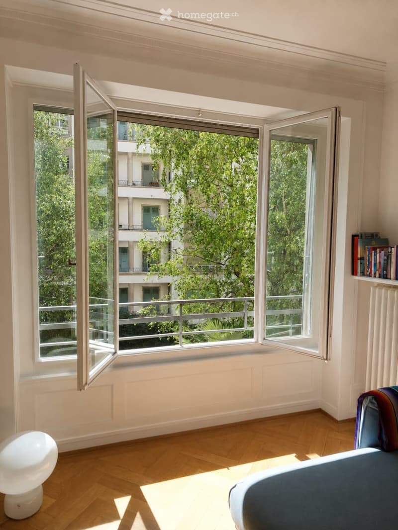 Bright room with wooden flooring, large open windows, a white stool, a blue sofa, and a bookshelf with books.