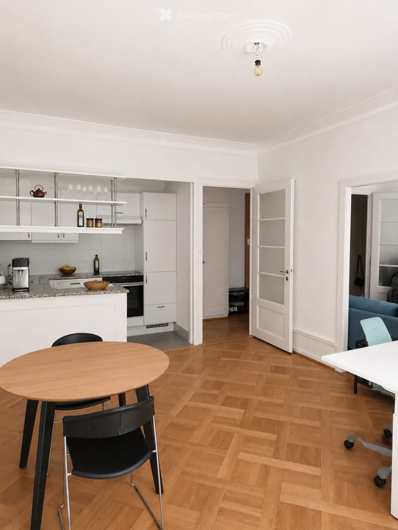 A modern, open-plan living and dining area with a round wooden dining table, black chairs, a white office chair, a blue sofa, and a kitchen area with white cabinets, a refrigerator, and an oven.