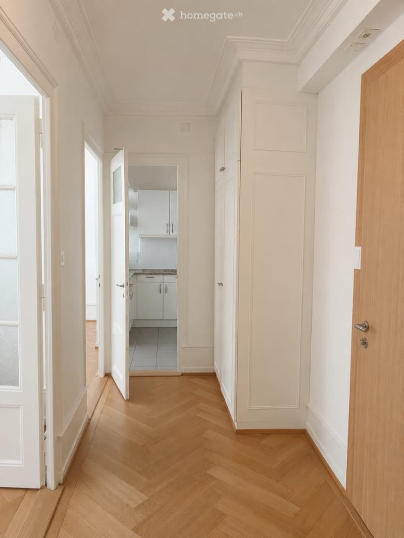 A well-lit hallway with wooden herringbone flooring, white walls, and decorative crown molding. Open doors reveal a modern kitchen with white cabinets and stainless steel appliances.
