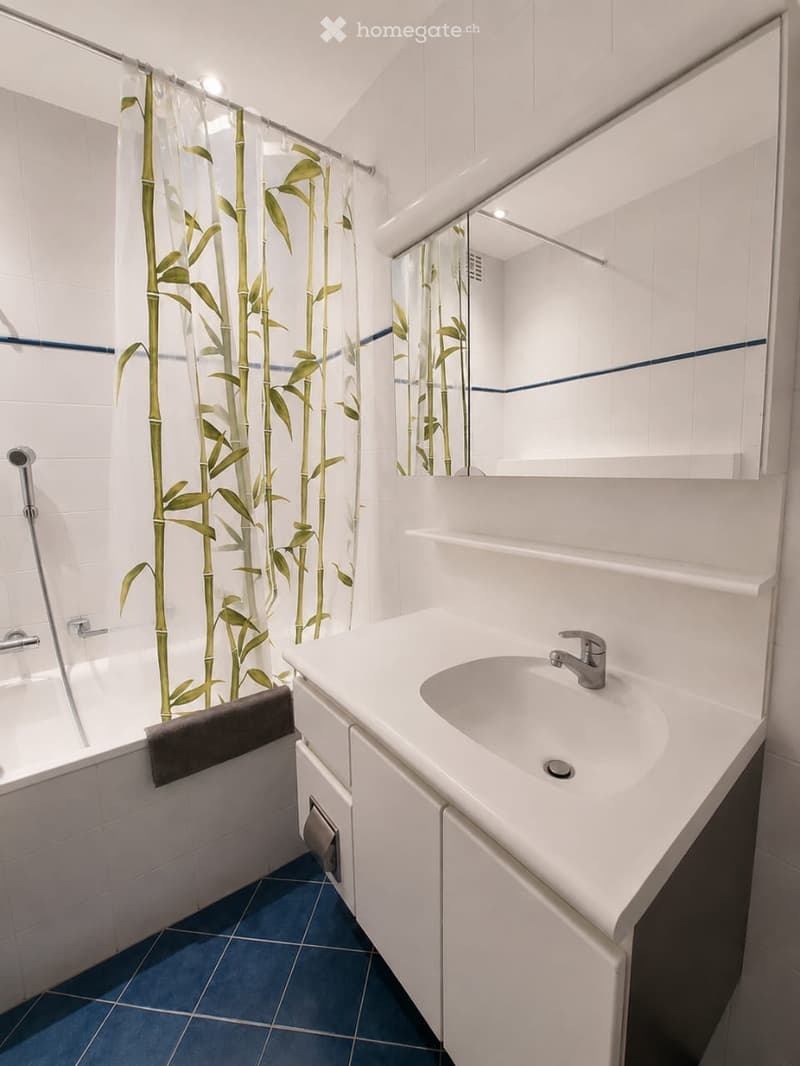 Modern bathroom with white tiles, blue accents, a bathtub, a sink with a faucet, a mirror, and a bamboo-patterned shower curtain.