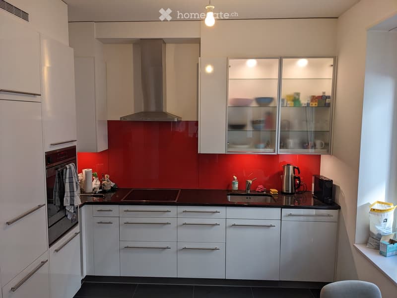 Modern kitchen with white cabinets, stainless steel appliances, and a red backsplash. Features an induction stove, sink, coffee maker, and various kitchen items on the counter.