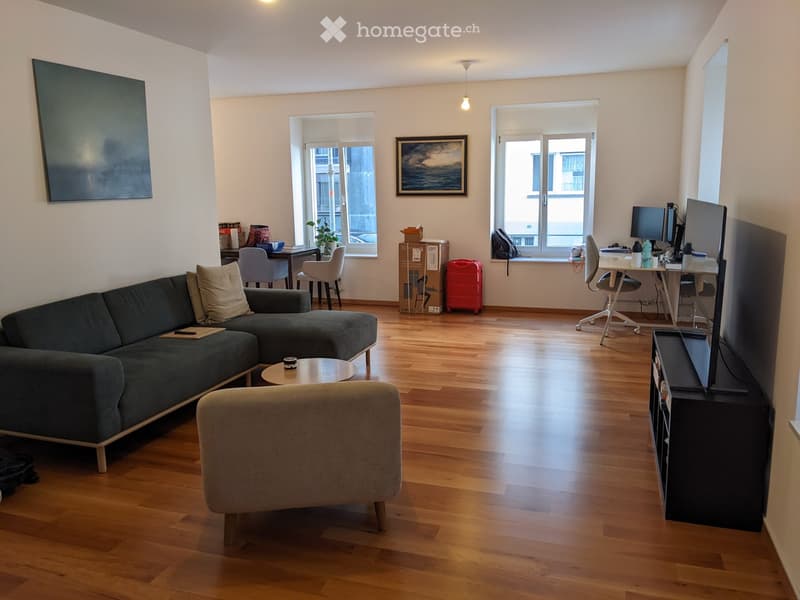 A modern living room with a dark green sectional sofa, a round coffee table, a grey armchair, a TV stand with a screen, a desk with a laptop, and a small table with a plant. The room has wooden flooring and large windows with blinds.