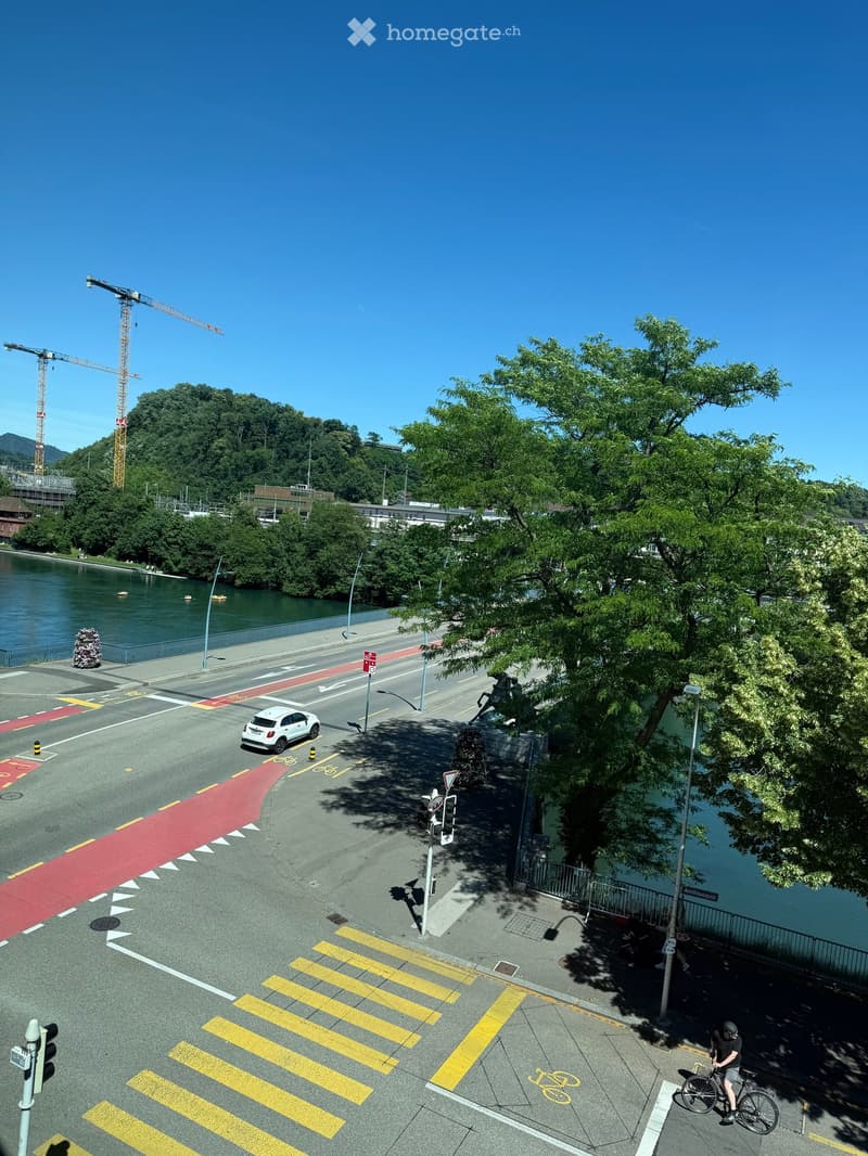 Aerial view of a riverside area with a road, pedestrian crossing, bicycle lane, and trees. A white car is visible on the road, and a person is riding a bicycle near the water.