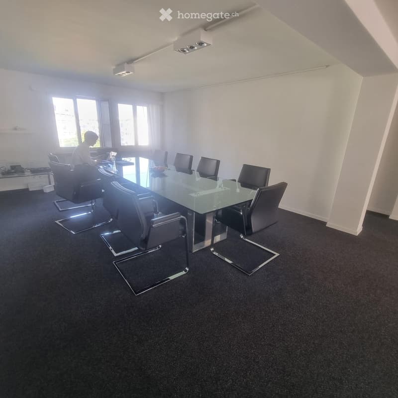 Conference room with a large glass table, black chairs, and a screen. The room has large windows and a carpeted floor.