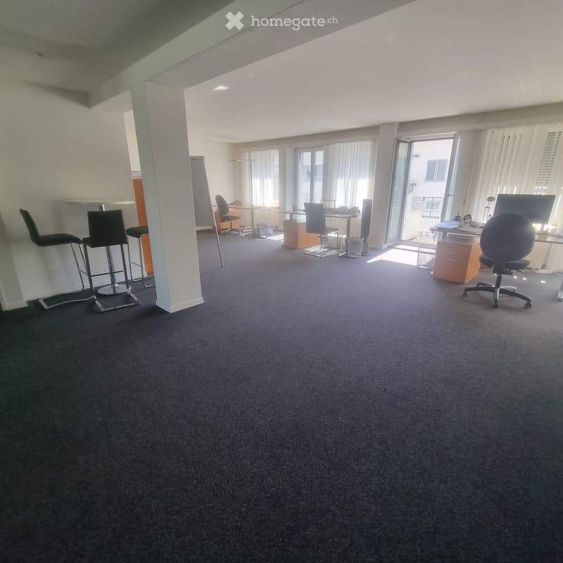 Office space with carpeted floor, large windows, and several desks with chairs, a screen, and a cabinet.
