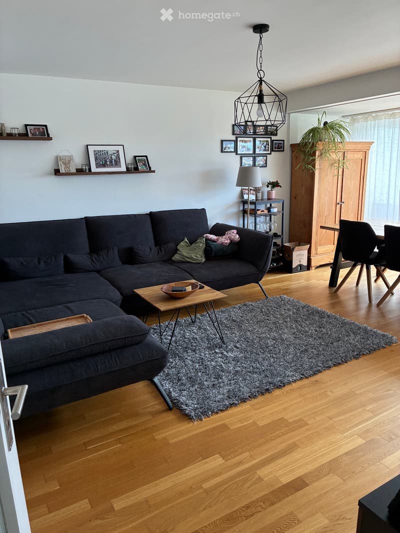 Modern living room with a dark grey sectional sofa, wooden coffee table, grey shag rug, and a wooden dining table with chairs. The room features wooden flooring, wall-mounted shelves with picture frames, and a hanging pendant light.