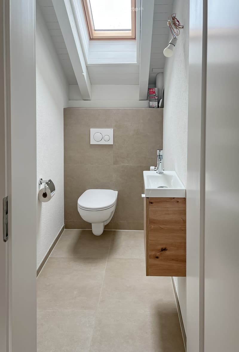 Modern, compact bathroom with a toilet, sink, and wooden cabinet. Features a skylight window, beige tiles, and a sleek design.
