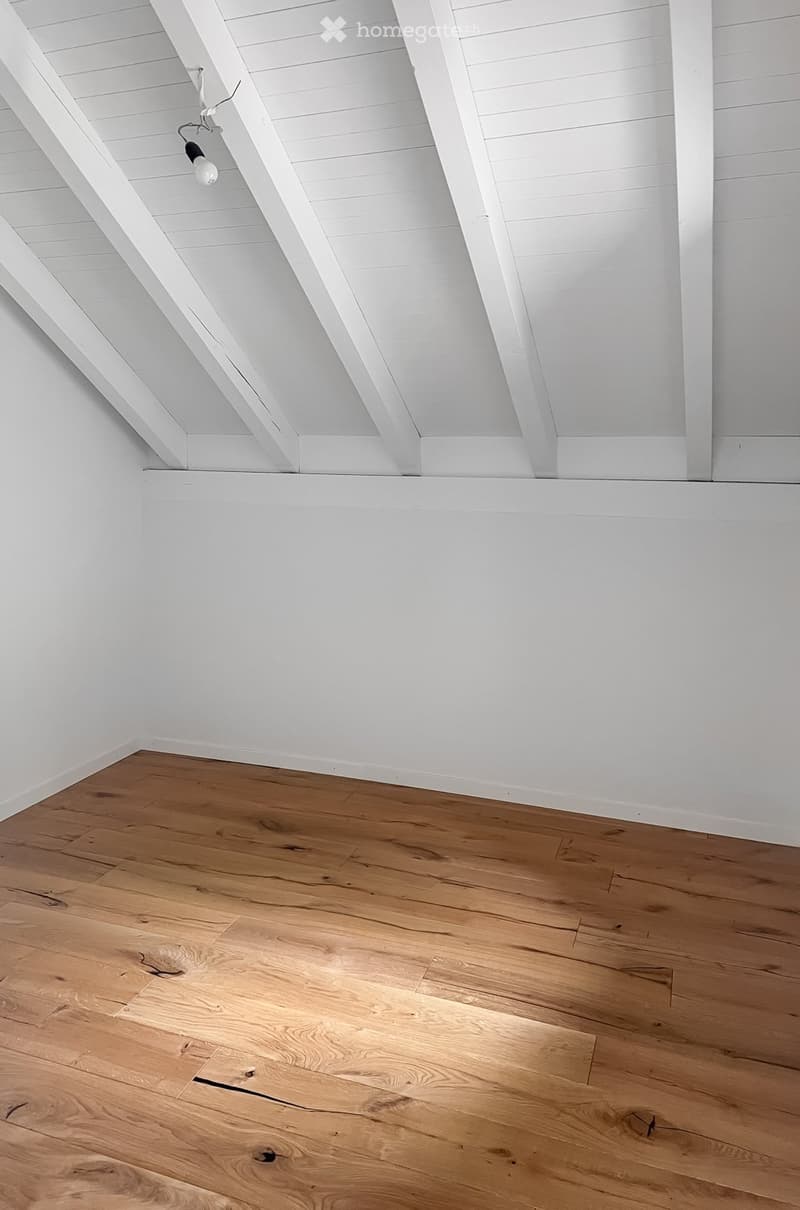 Empty room with wooden flooring and white walls, featuring exposed wooden beams on the ceiling and a single light fixture.