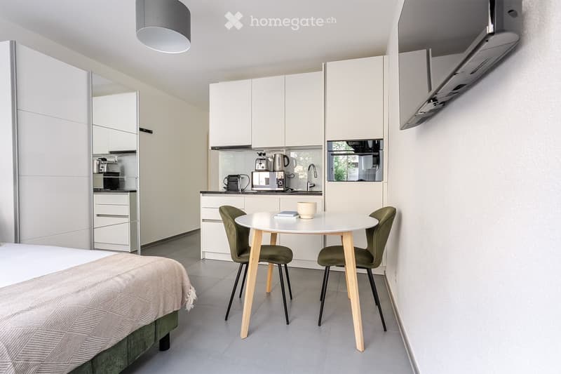 A modern, compact studio apartment featuring a bed, a small dining table with two chairs, a kitchen area with cabinets, a sink, and various appliances including a coffee maker, microwave, and oven. The space is well-lit with a hanging lamp and has a sleek