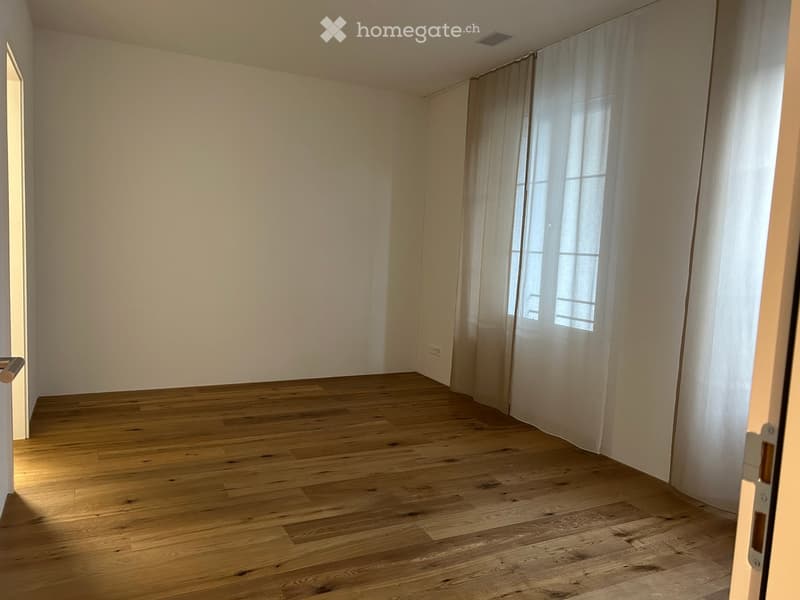 Empty room with wooden flooring, white walls, and a window with sheer curtains.
