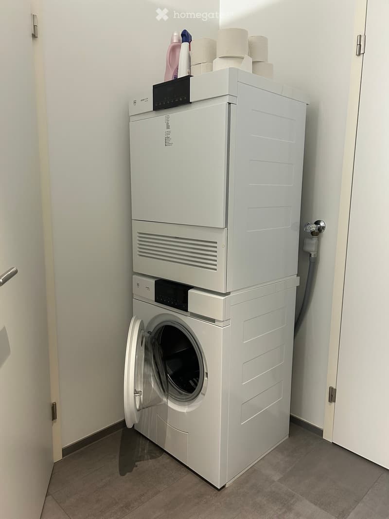 A compact bathroom with a stacked washing machine and dishwasher unit, toilet paper on top, and a tiled floor.
