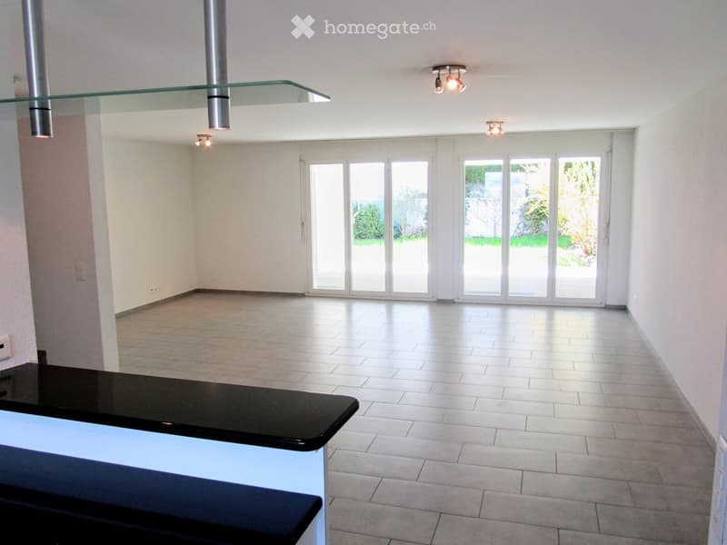 Empty room with white walls, tiled floor, large sliding glass doors leading to a garden, and a black countertop in the foreground.