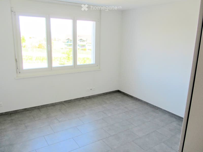 Empty room with white walls, tiled floor, and large windows offering a view of the outside area.