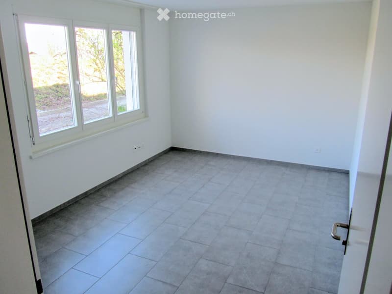 Empty room with white walls, tiled floor, and large windows with a view of greenery outside.
