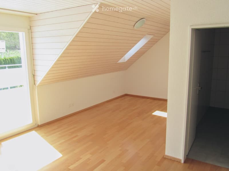 Empty room with wooden flooring, white walls, and a slanted wooden ceiling. Features a large window and a door leading to another room.