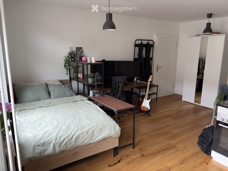A modern, well-lit bedroom with a wooden bed, a desk, a guitar, and a small table. The room features a large window with a view, a hanging pendant light, and a small plant on the windowsill.