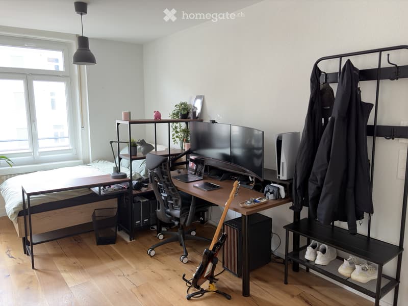 Modern, multi-functional room with a lofted sleeping area, a desk, and storage shelves. Features a sleek black chair, a wooden table, a guitar, and a modern lamp on the floor.