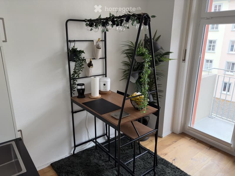 A modern, compact workspace with a wooden desk, black metal shelving unit, laptop, bowl of fruit, and decorative plants. The room features large windows with a view of the city and a balcony accessible through the window.