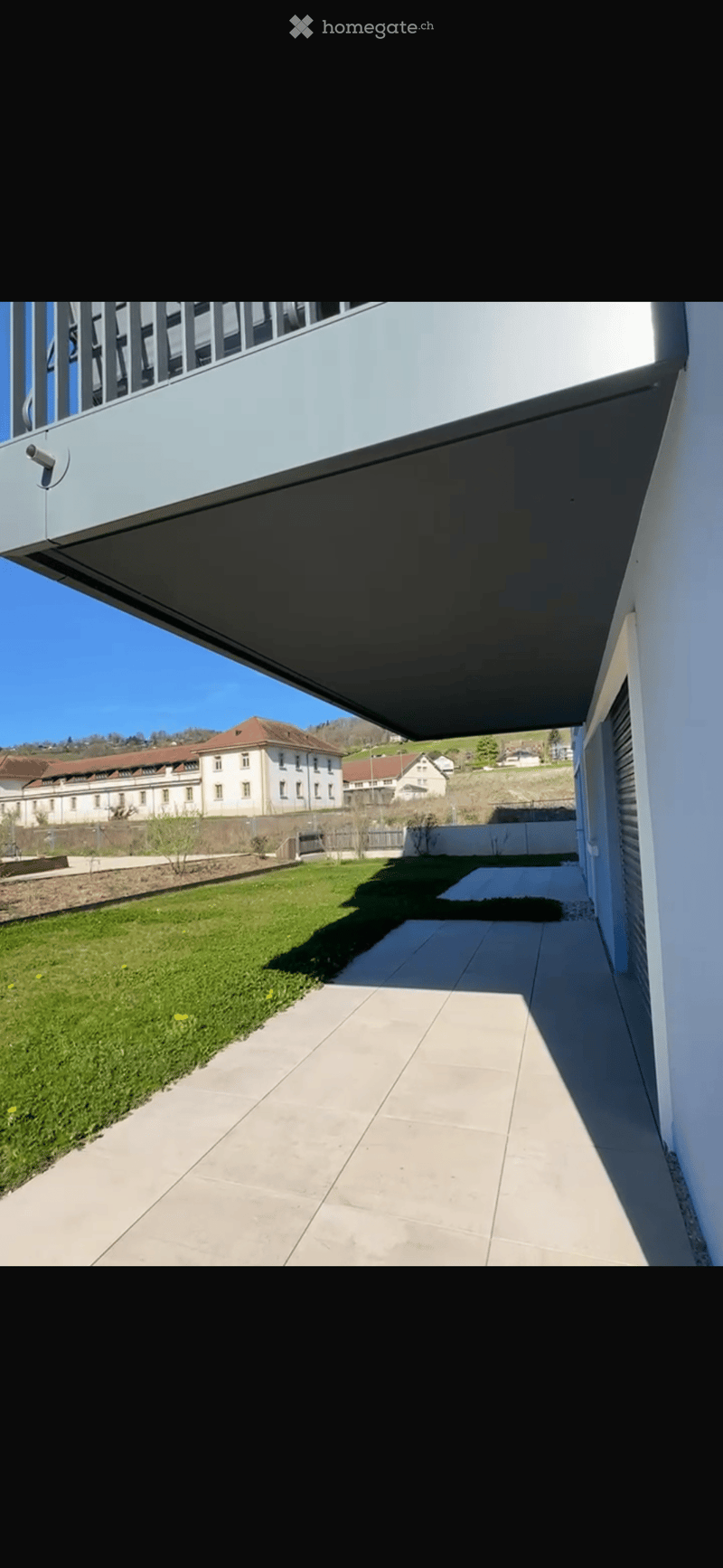 A modern outdoor area with a paved walkway, grassy lawn, and a view of a distant building complex.