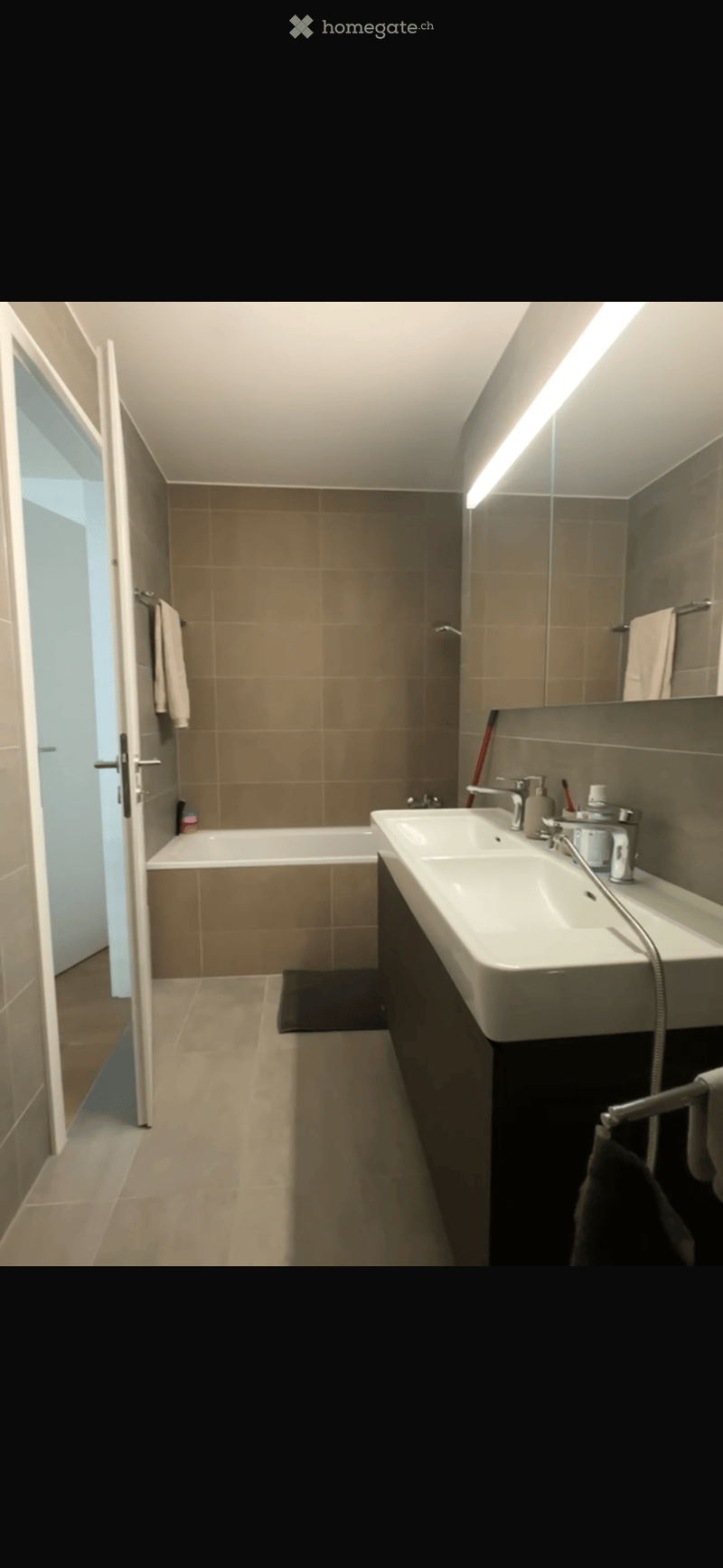 Modern bathroom with a bathtub, double sink, large mirror, and towel rack. The space features beige tiles and a sleek design.