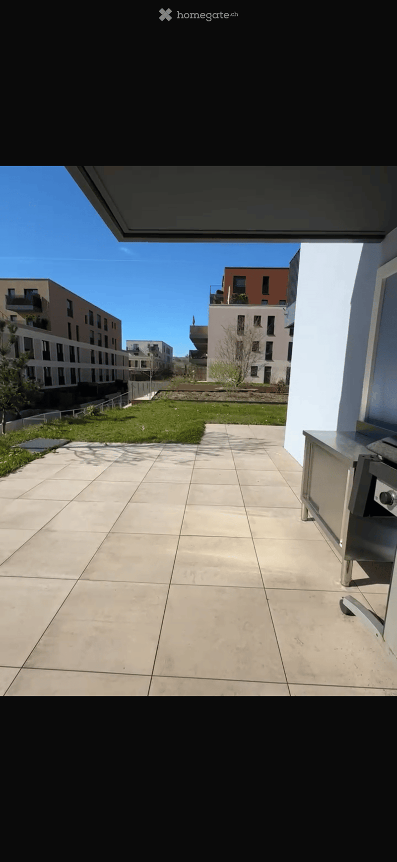 Spacious balcony with tiled floor, modern stainless steel railing, and a view of a landscaped area with grass and modern apartment buildings.