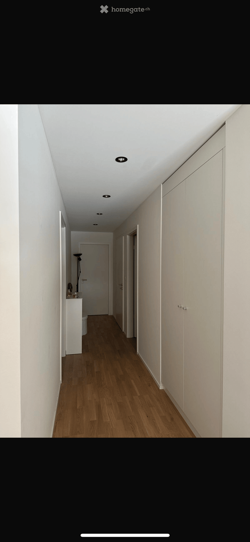 A long, narrow hallway with light wooden flooring, white walls, and recessed ceiling lighting. Features built-in white cabinetry on one side and a small table with a lamp on the other side.