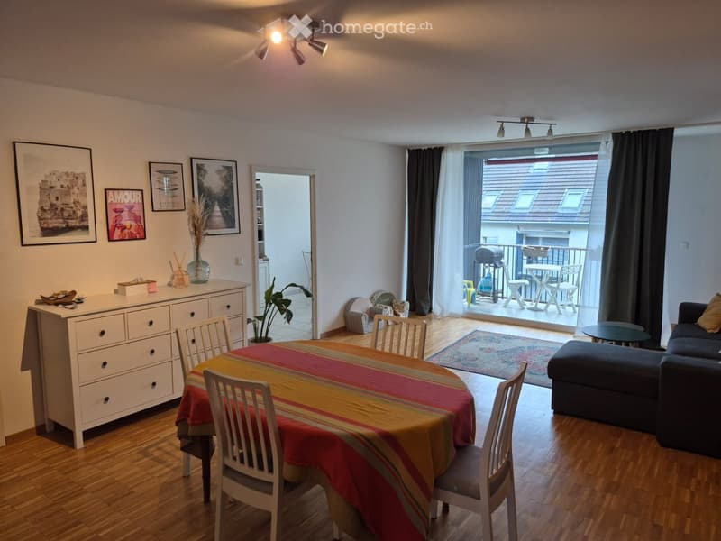 Bright living and dining area with hardwood floors, large windows, and a balcony. Features a sofa, ottoman, coffee table, dining table with chairs, cabinet, and decorative items.