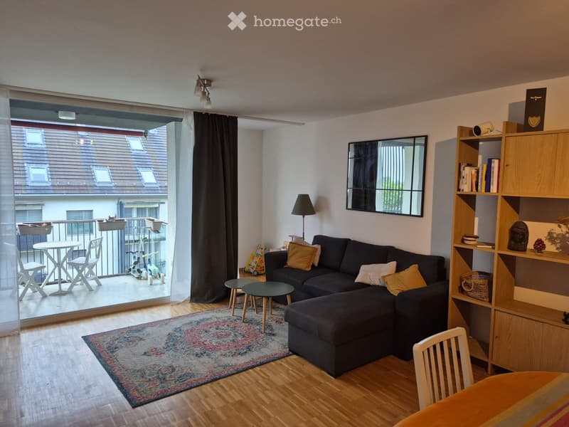 Modern living room with a dark sectional sofa, wooden coffee table, side table, floor lamp, bookshelf, and a balcony with outdoor furniture.